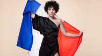 Barbara Pravi in a black dress. She is holding the Franch flag behind her.