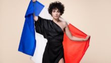 Barbara Pravi in a black dress. She is holding the Franch flag behind her.