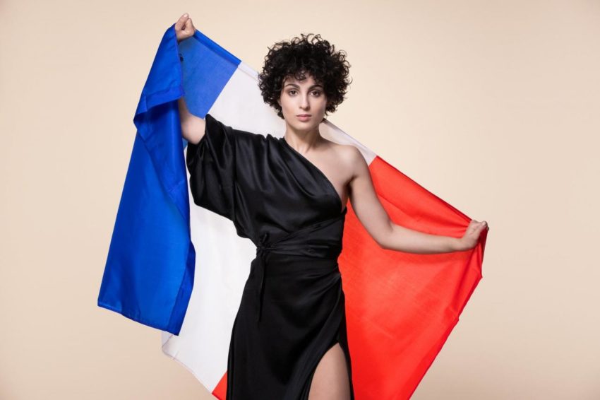 Barbara Pravi in a black dress. She is holding the Franch flag behind her.