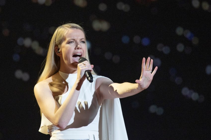 Ana Soklič performs in a white gown. She is holding out her left arm in front of her.